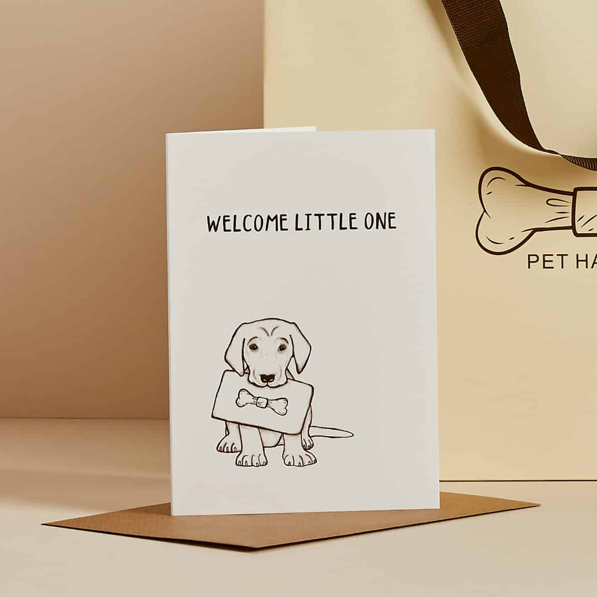 White greeting card with illustrated puppy holding sign, 'Welcome Little One' text, kraft envelope