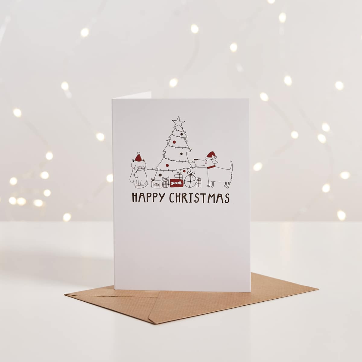 White Christmas card with illustrated cat and dog decorating a tree, 'Happy Christmas' text, kraft envelope