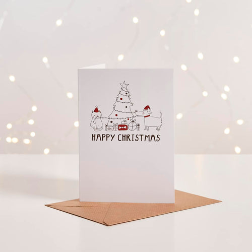 White Christmas card with illustrated cat and dog decorating a tree, 'Happy Christmas' text, kraft envelope