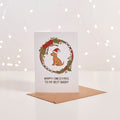 White Christmas card with illustrated dog in Santa hat inside a wreath, 'Happy Christmas to my best buddy' text, kraft envelope