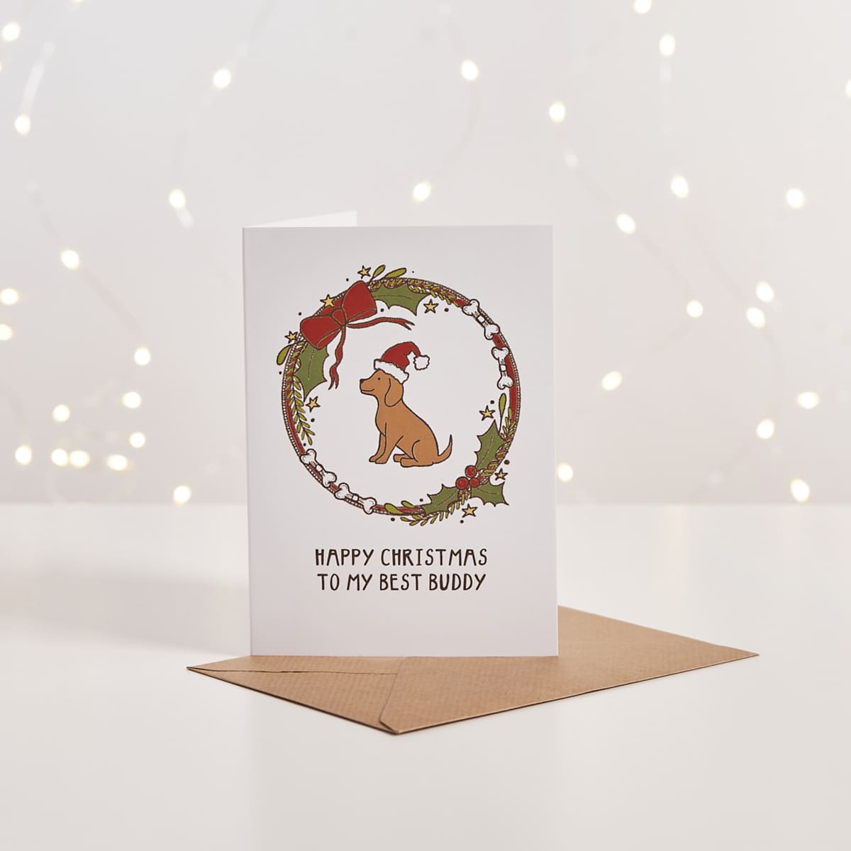 White Christmas card with illustrated dog in Santa hat inside a wreath, 'Happy Christmas to my best buddy' text, kraft envelope